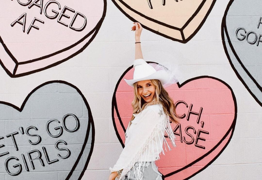 Bride dressed in white fringe jacket, white cowboy hat, and blue jeans (resembling the disco cowgirl style), is raising her arm up pointing to the sky with a big smile on her face in excitement while standing in front of a wall painted with a colorful candy hearts mural.